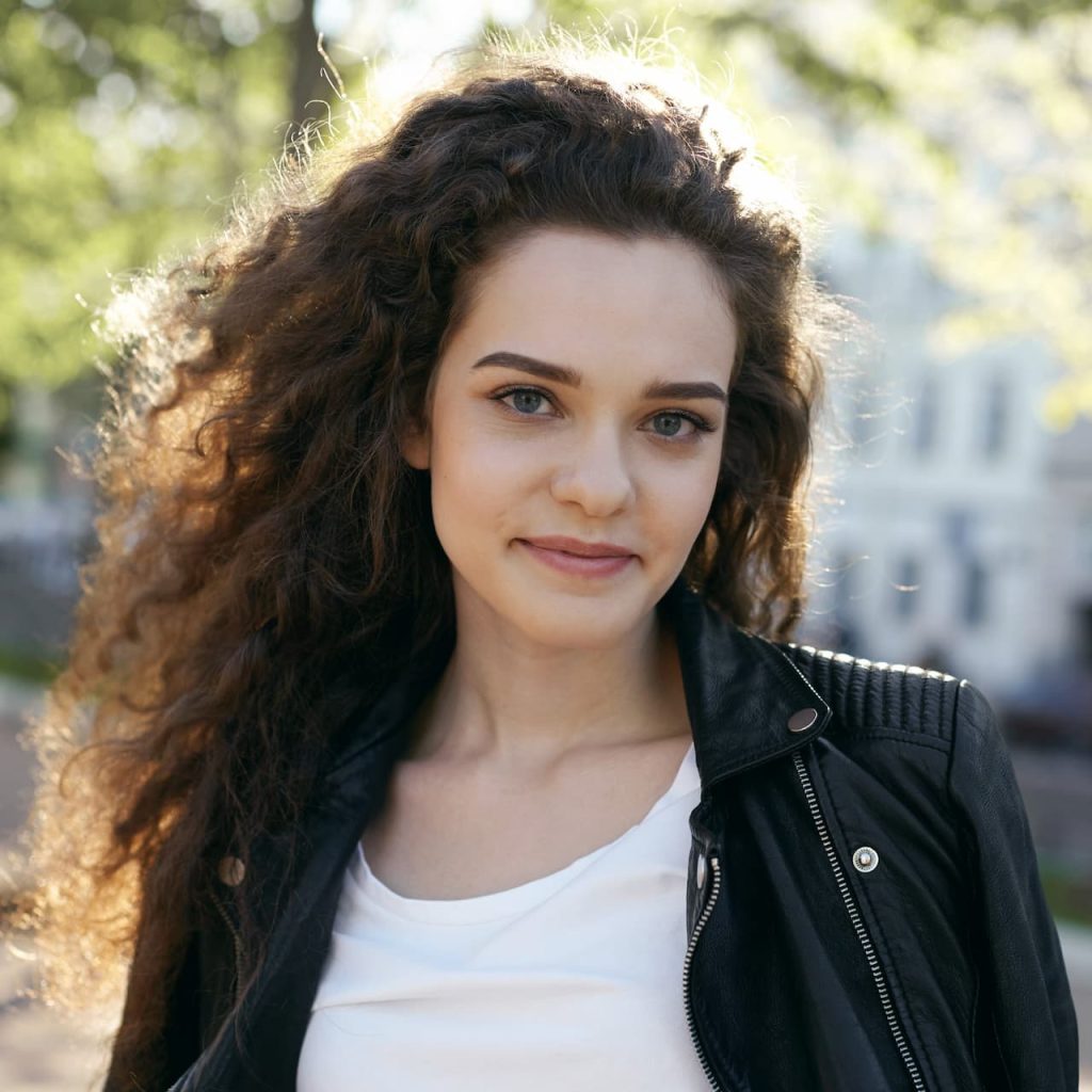 A smiling young woman with curly hair, a white t-shirt, and a black leather jacket