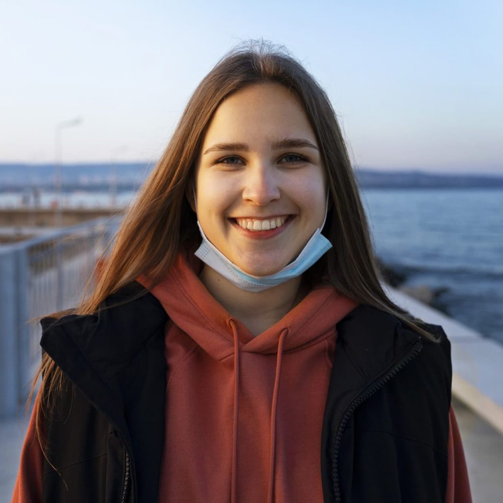 A smiling young woman with long hair and a mask under her chin near the water